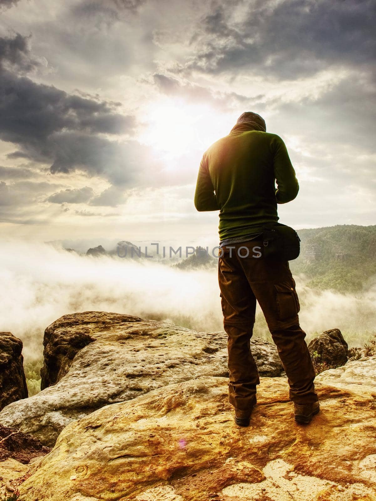 Landscape photograper with camera ready in hand. Man climbed up on exposed rock for fall photos with his digital camera. Cliff above misty autumn landscape.