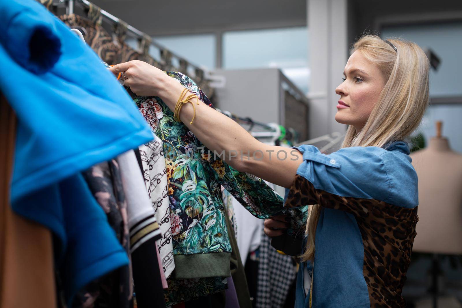 A woman looks at the new clothes hanging on the hangers. A blonde woman with long hair looks for the right blouse for her.