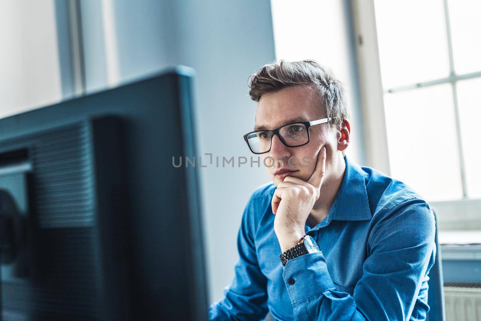 Handsome Young Businessman Wearing Eyeglasses Sitting at his Table Inside the Office, Looking at the Report on his Computer Screen