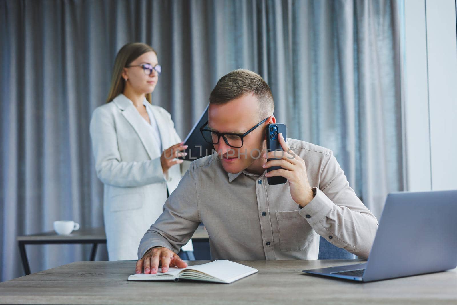 A male manager is talking on a mobile phone while sitting at a table with a computer and a notepad. Working atmosphere in an office with large windows