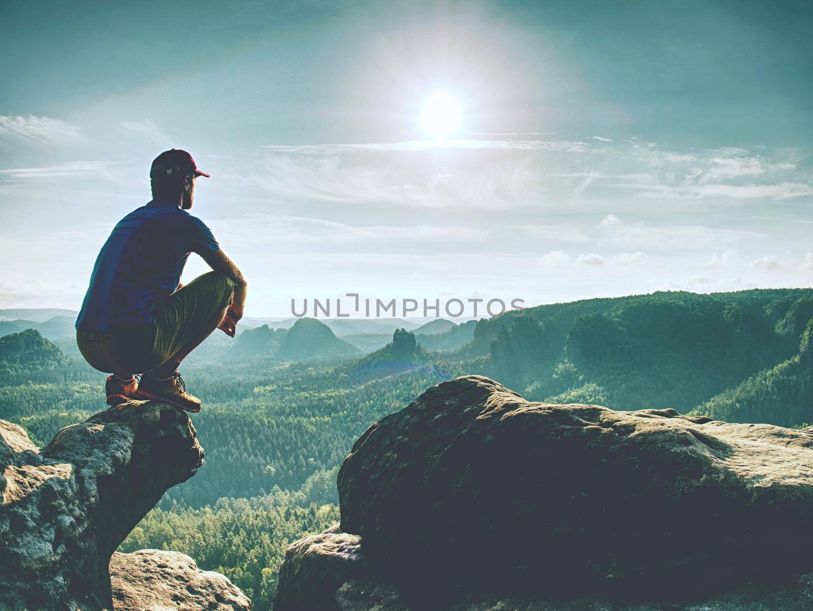 Sit on the edge. Man in blue yellow sports clothes  sit on sharp cliff and enjoying far view. View into misty hilly valley bellow