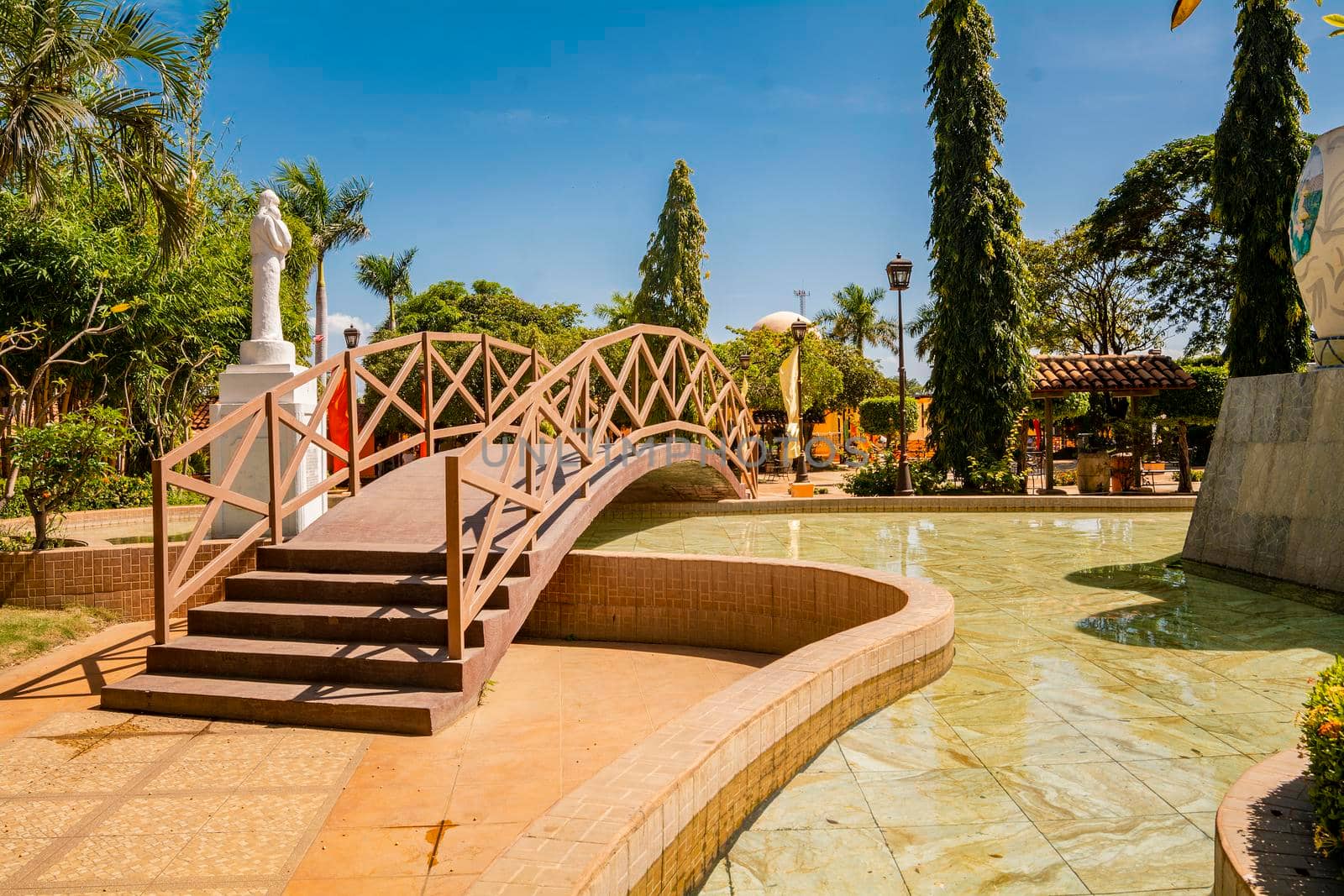 View of a calm park with a small wooden bridge over a water fountain. Nagarote central park.  Image of a nice and relaxed park with a water fountain surrounded by trees. Traditional park of Nagarote, Nicaragua by isaiphoto