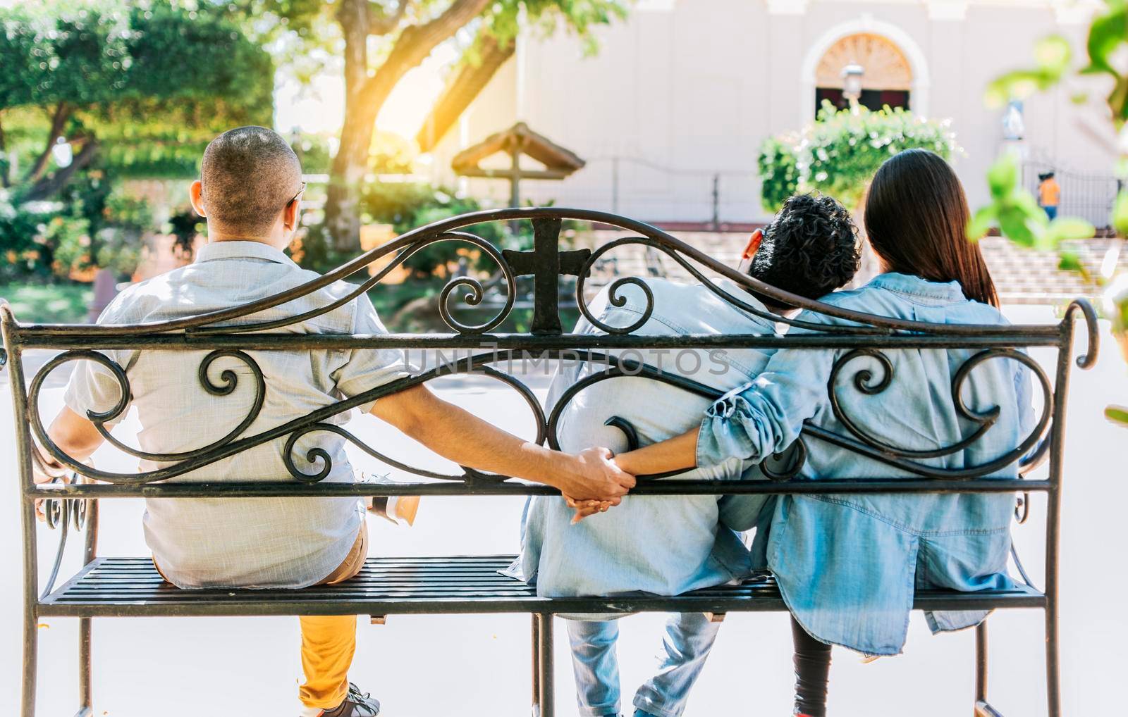 Unfaithful woman sitting holding hands with another man while boyfriend hugs her. Unfaithful couple holding hands on a park bench. Concept of love triangle and infidelity by isaiphoto