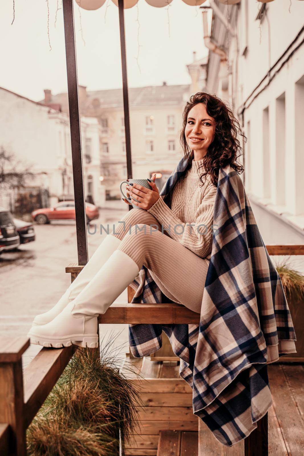 A middle-aged woman in a beige sweater with a blue mug in her hands is in a street cafe on the veranda by Matiunina