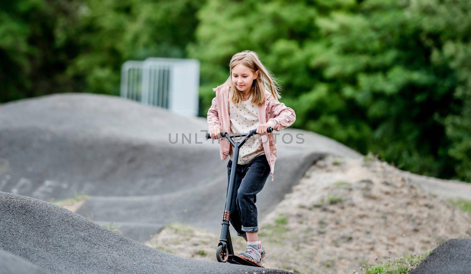 Preteen girl riding scooter in city park at spring time. Pretty child kid having fun with vehicle outdoors