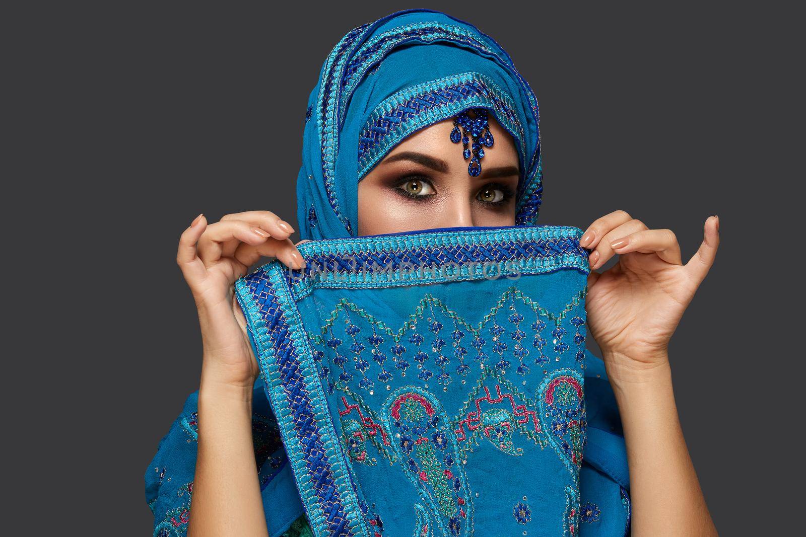 Close-up portrait of a white-skinned girl with beautiful smoky eyes wearing an elegant blue hijab decorated with sequins and jewelry. She is covering her face with a shawl and looking at the camera on a dark background. Human emotions, facial expression concept. Trendy colors. Arabic style.