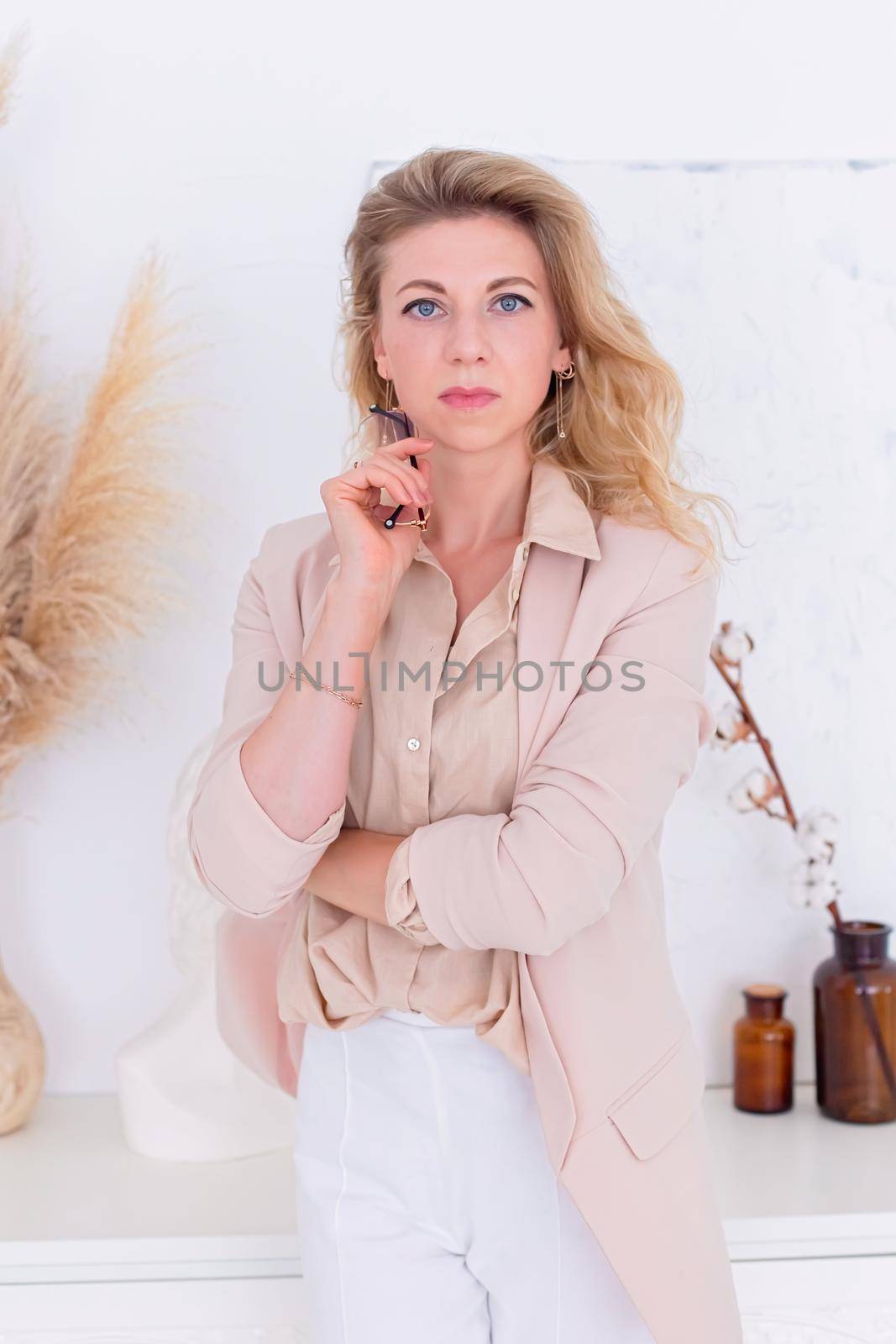 Portrait of beautiful woman with blond hair, stands in a light pink jacket in the interior of a white room, look to the camera, hold glasses . Copy space. Vertical