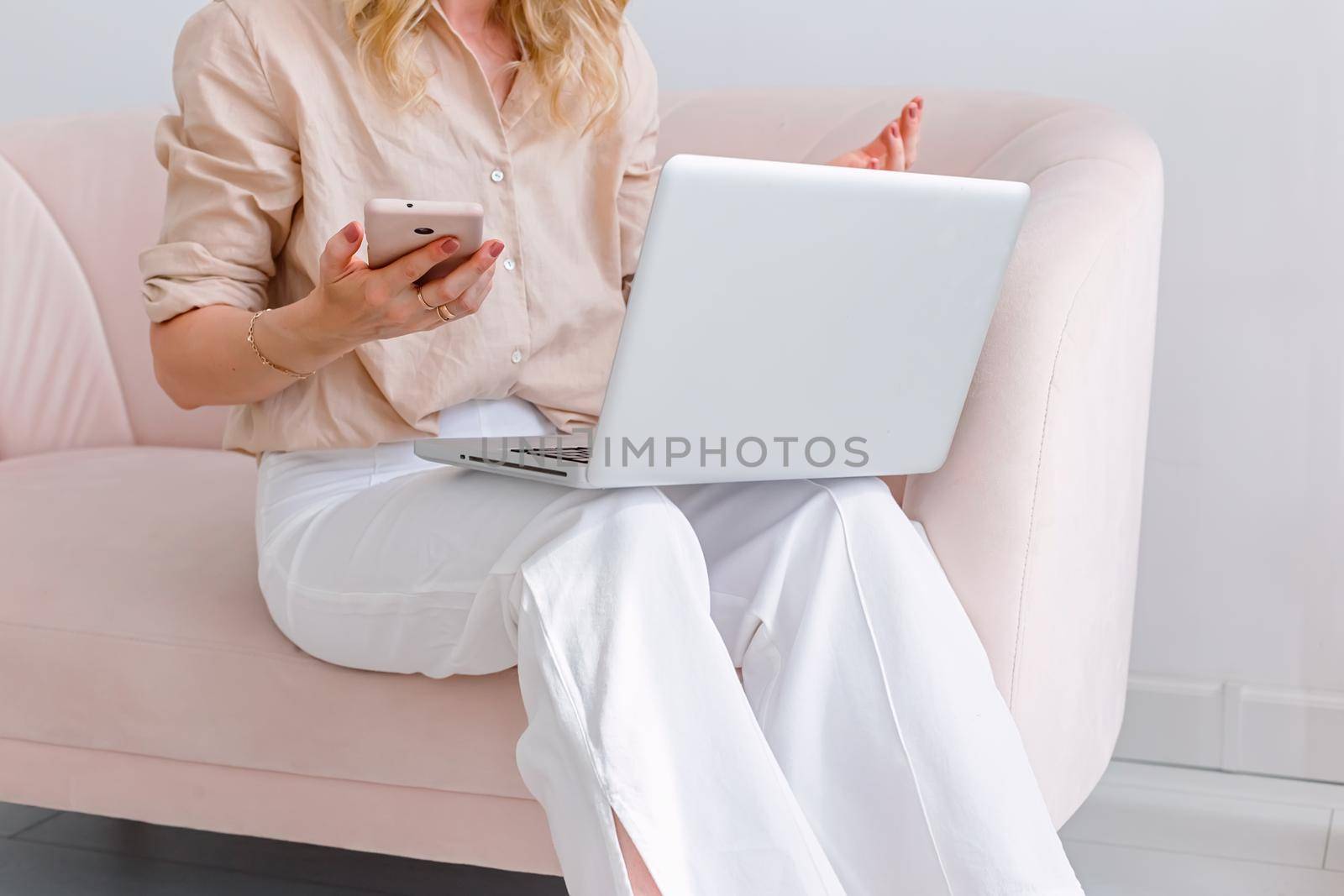 A business woman in light clothes, sits on a light pink sofa, with a gray open laptop on her knees, holds a pink smartphone in her hand, close-up