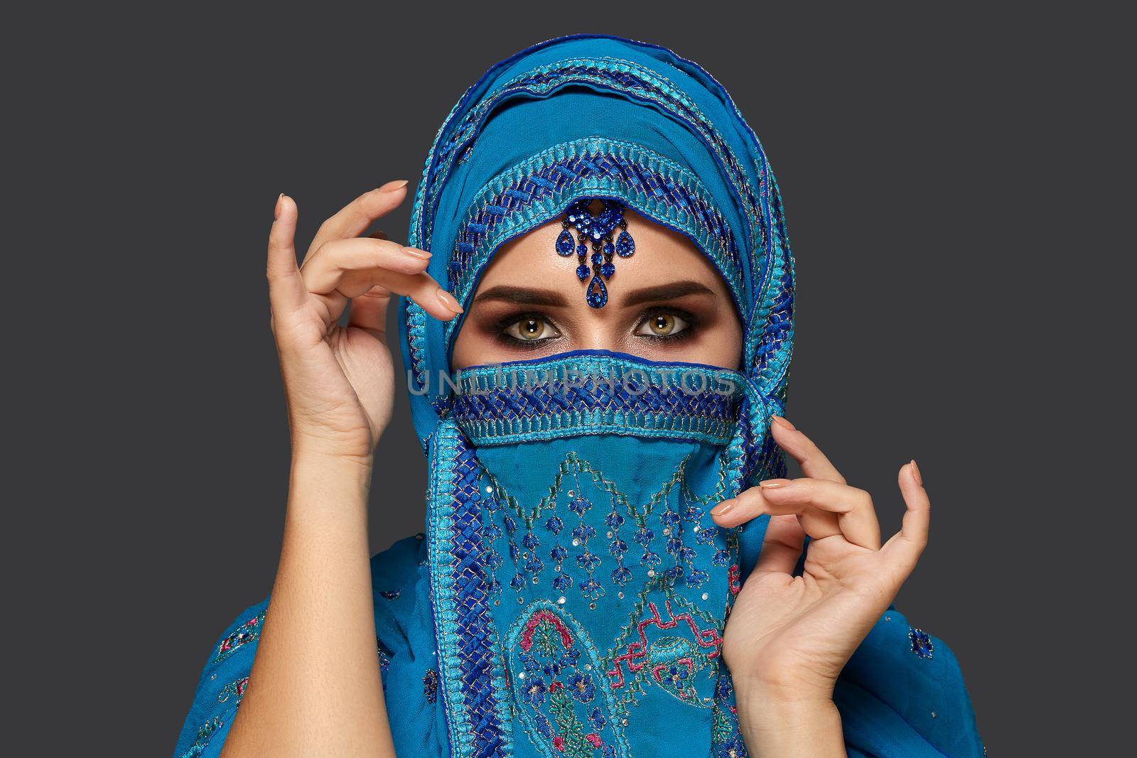 Close-up portrait of a lovely young woman with beautiful smoky eyes wearing a blue hijab decorated with sequins and jewelry. She is gesticulating and looking at the camera on a dark background. Human emotions, facial expression concept. Trendy colors. Arabic style.