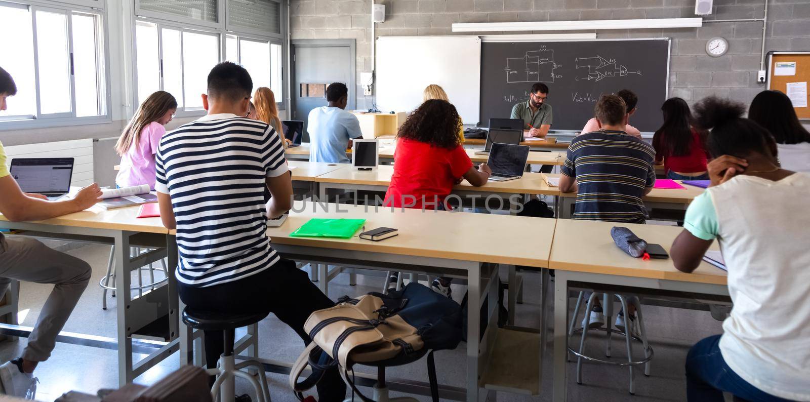 Rear view of group of multiracial high school students in class using laptops while teacher marks exams. by Hoverstock