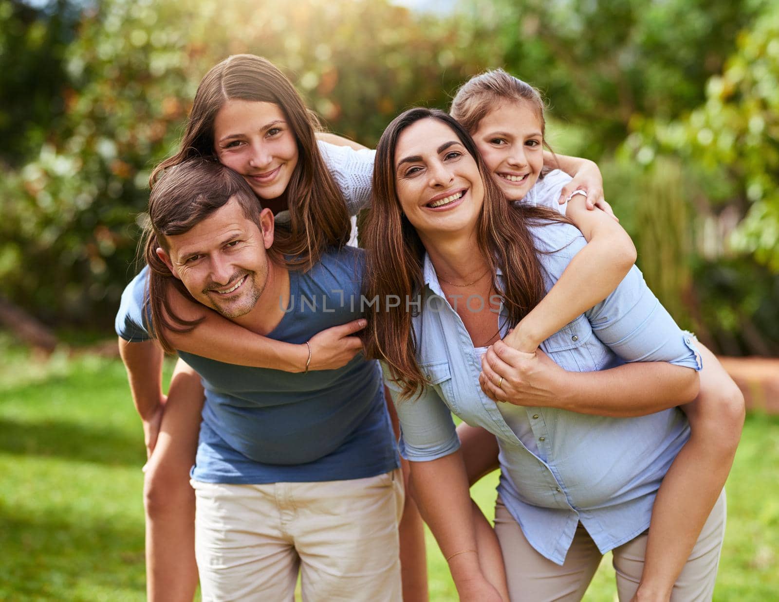 Lets take a ride. Portrait of a cheerful young mother and father giving their daughters a piggyback ride outside in a park during the day