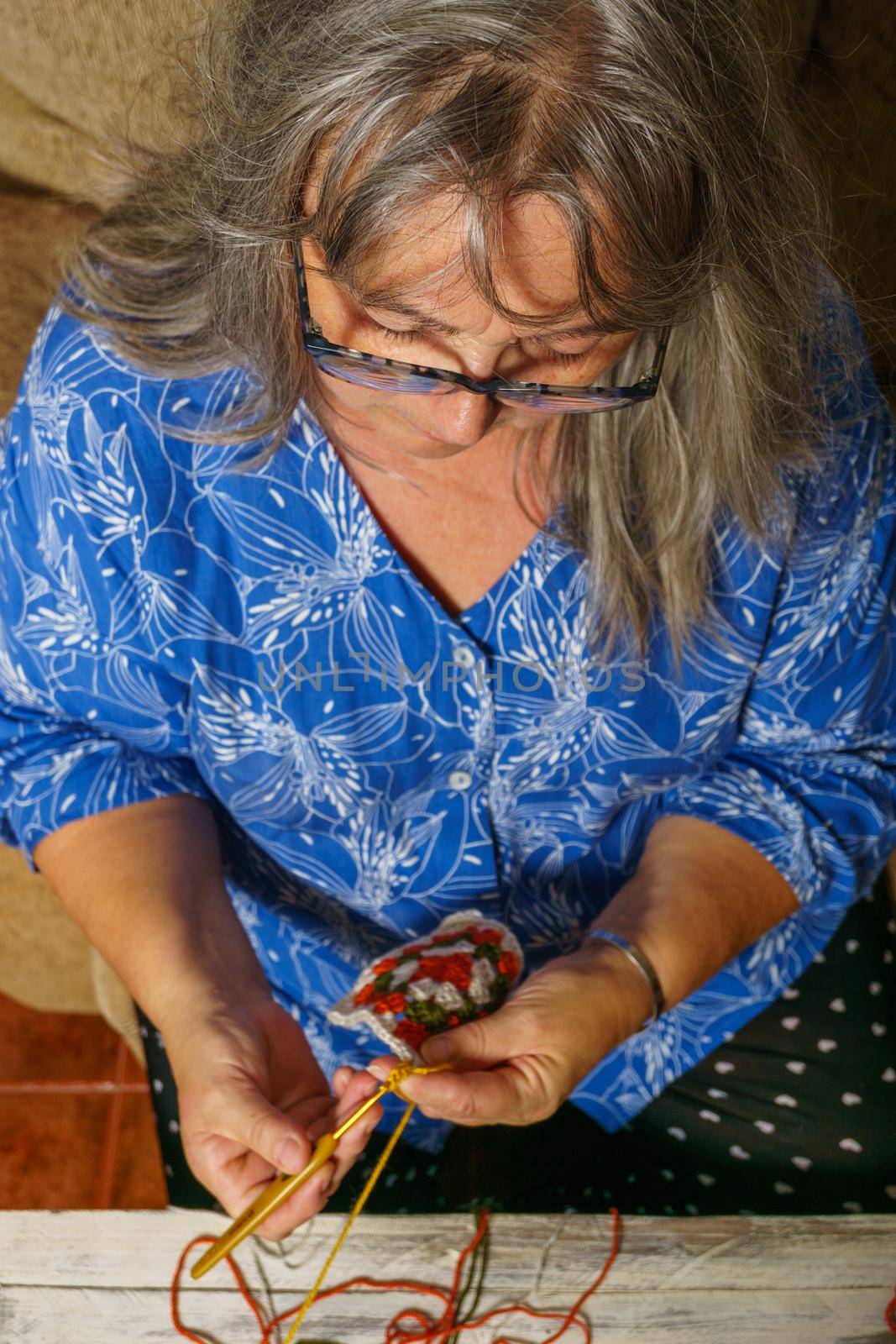 older woman with white hair and glasses crocheting on the sofa at home