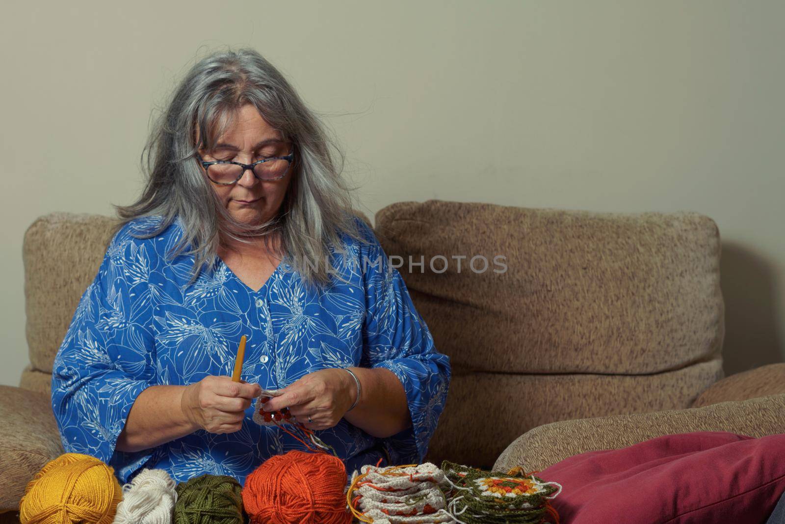 older woman with white hair and glasses crocheting on the sofa at home