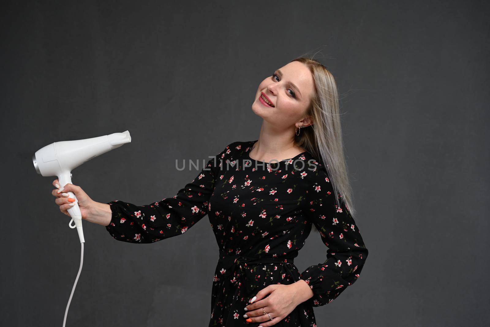 Portrait of a beautiful young blonde woman with long straight hair on a gray background with a hair dryer in her hands