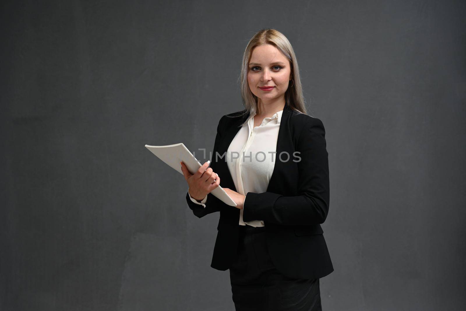 Portrait of a confident young businesswoman wearing formal suit standing isolated over gray background, arms folded by chichaevstudio