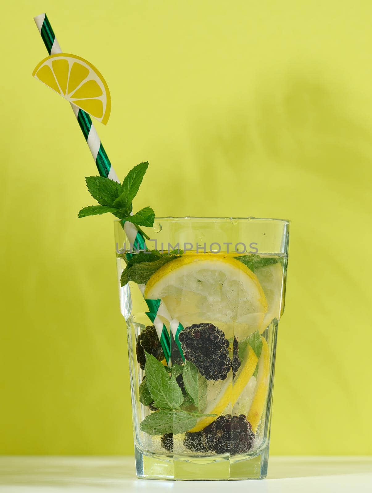 Transparent glass with lemonade, mint leaves, lemon slices and blackberries in the middle. Green background