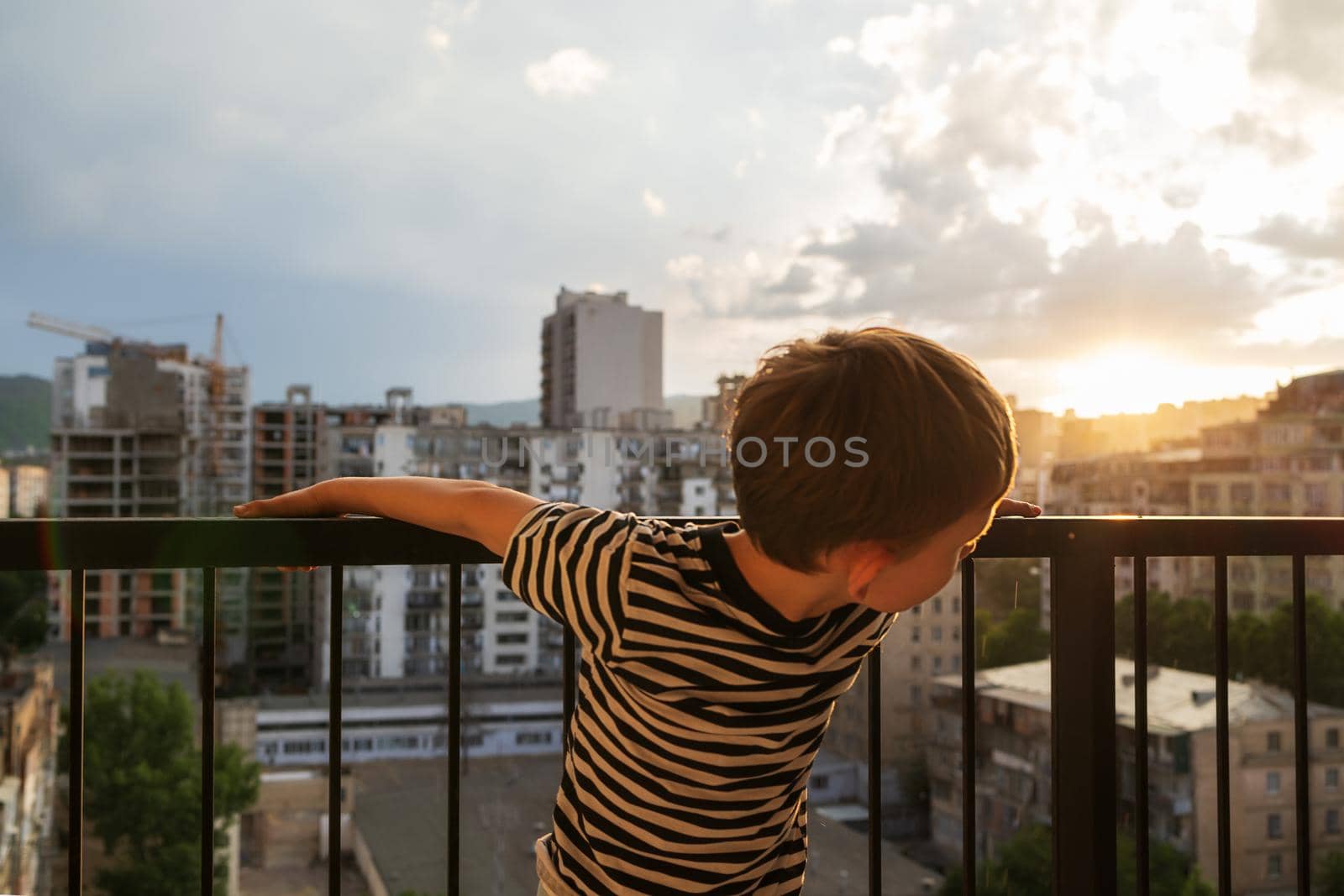 A 6-year-old boy climbs the dangerous high railing of a balcony unsupervised. City in the evening at sunset. Child at home alone. Risky child behavior. Deadly accidents. Altitude, danger