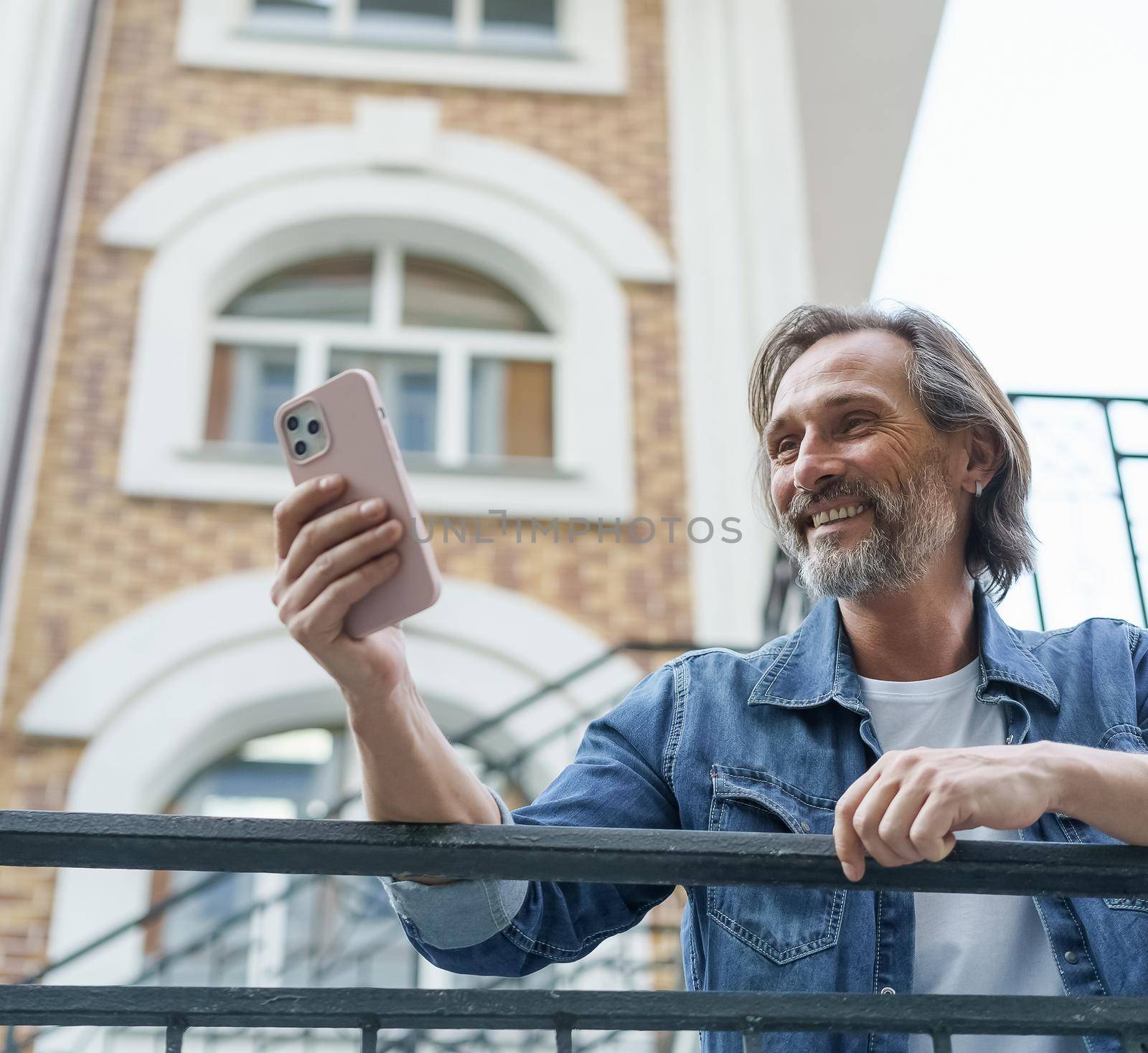 Happy middle aged grey bearded man laughing reading text message or funny social media post using smartphone standing in old town wearing casual. Travel concept.