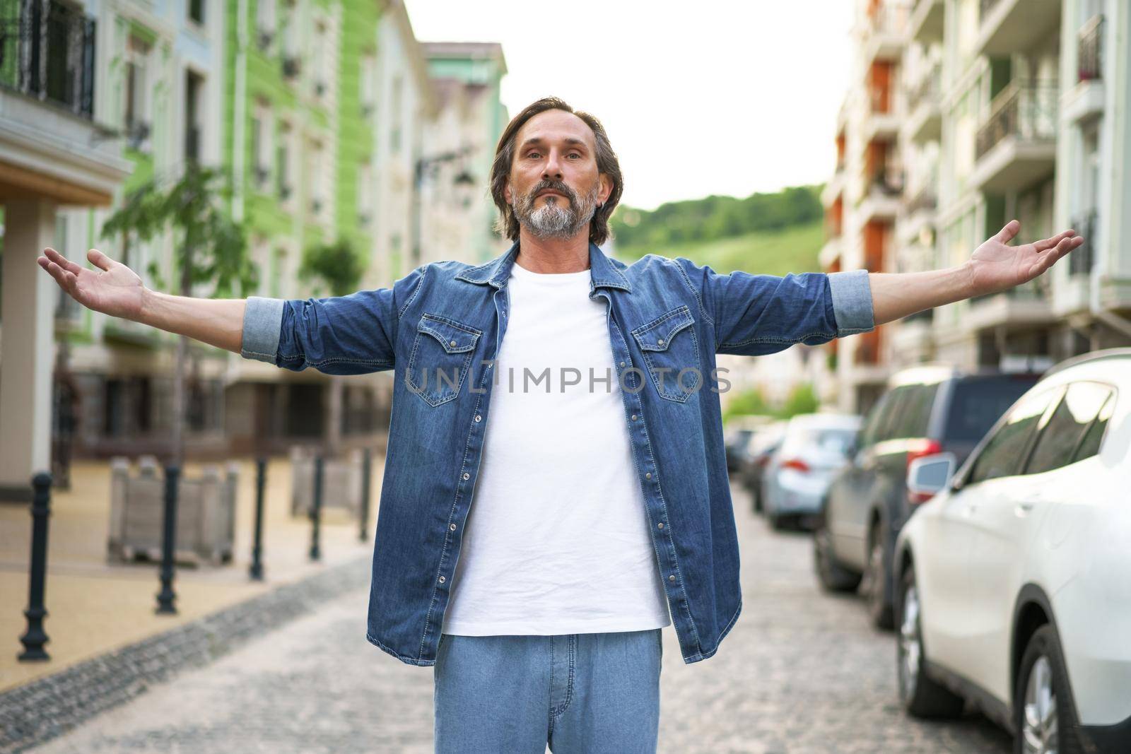 Happy senior man thanks God standing outdoors with hands spread wide open wearing jeans shirt with white t-shirt under. Mid Aged man glad that life is beautiful standing in old town street.
