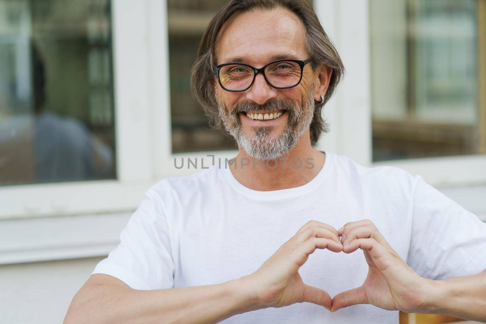 Gesturing heart, love, happy mature man in glasses smile standing outdoors wearing white t-shirt. Happy mature man in eye glasses and looking at camera sending love outdoor on a summer day.