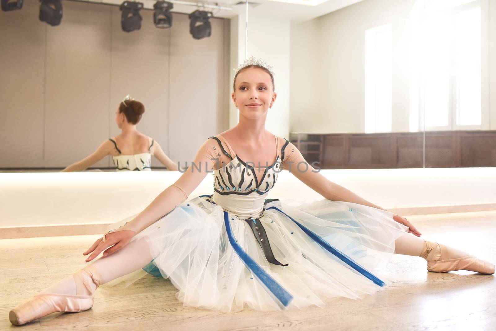 A young ballerina practicing ballet poses sitting against the mirror in the dance studio. by Nickstock