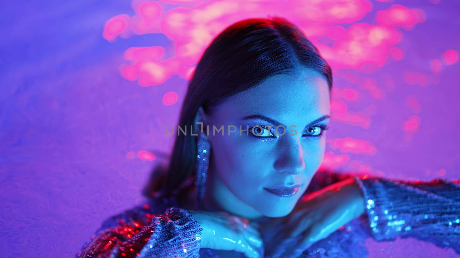 Beautiful woman posing in pool water under neon color light. Party, attractive chic in shining dress enjoying night time. Rich lifestyle, luxury life, influential people. High quality photo