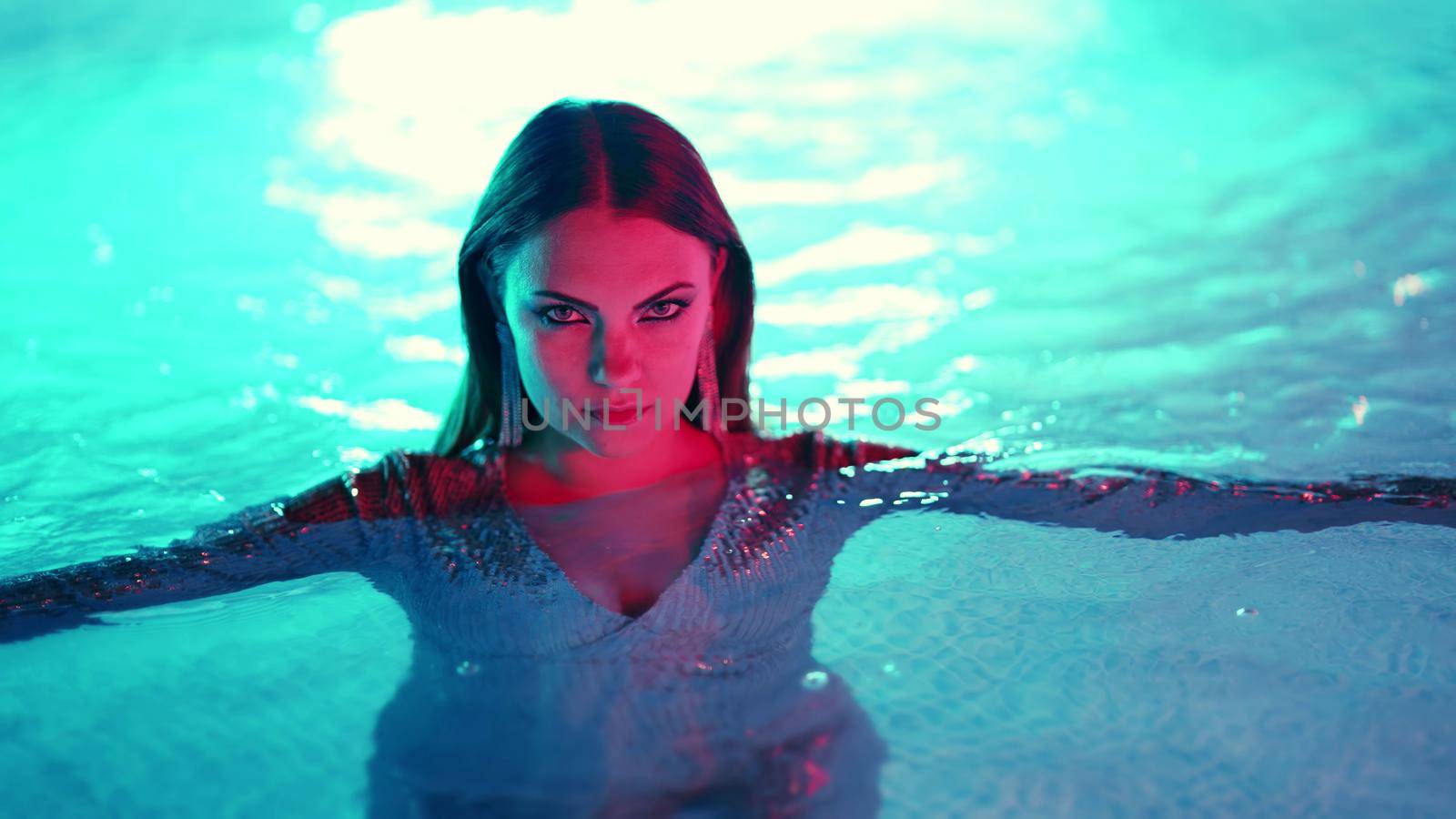 Beautiful woman posing in pool water under neon color light. Party, attractive chic in shining dress enjoying night time. Rich lifestyle, luxury life, influential people. High quality photo