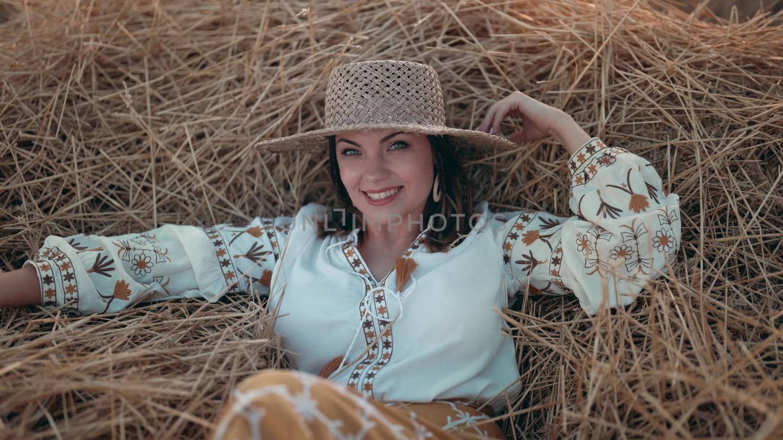 Pretty woman in straw hat and embroidered blouse smiling lying on hay in countryside at sunset. Rural nature, haystack, vacation, relax and harvest concept. High quality photo