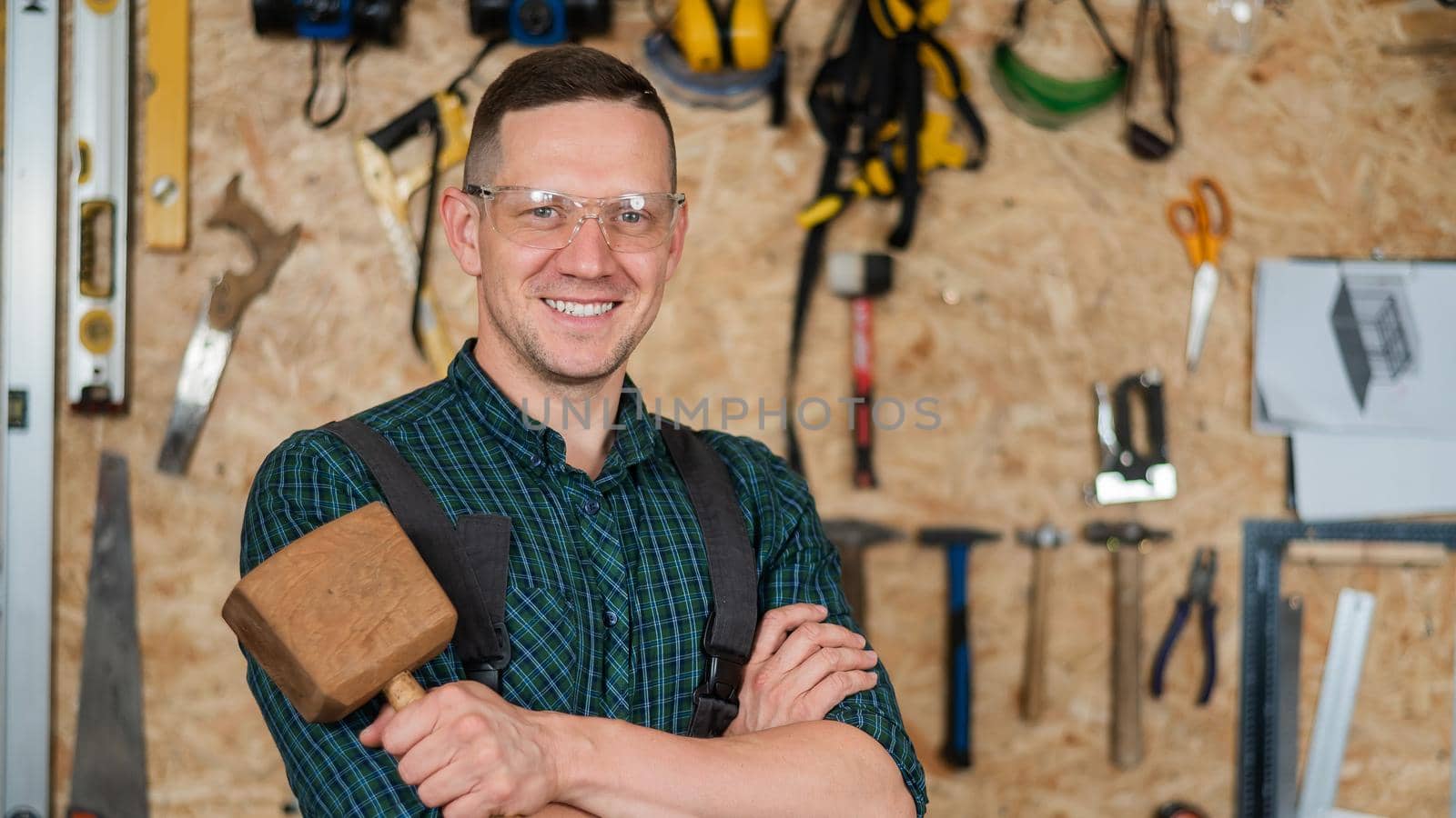 Portrait of a carpenter in goggles and overalls holding a wooden hammer in the workshop against the background of a wall with tools. by mrwed54