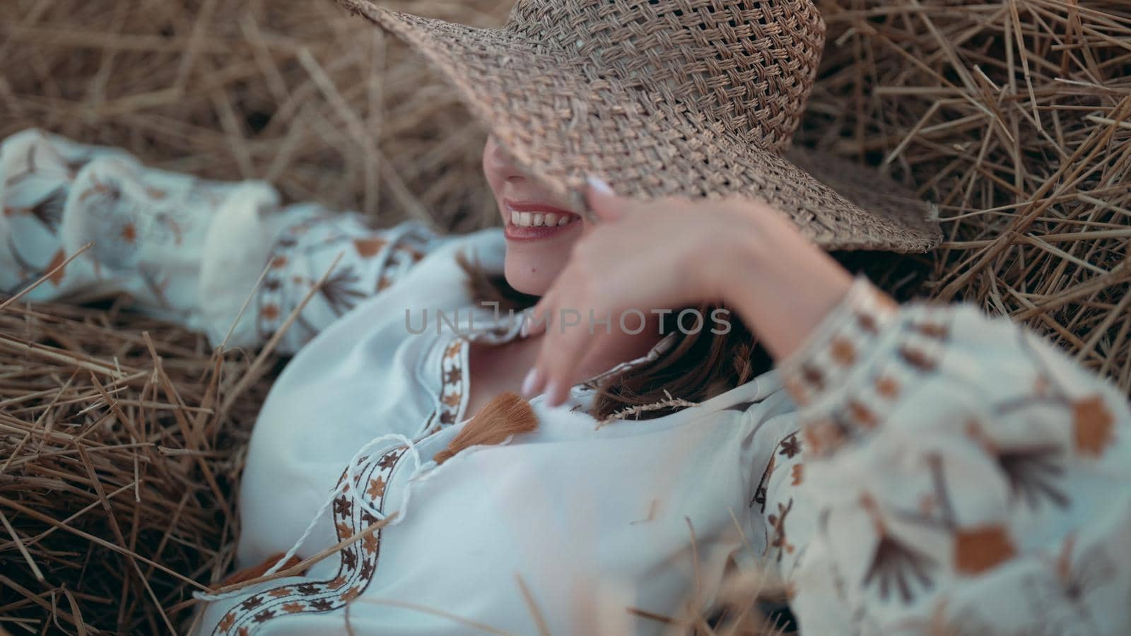 Pretty woman in straw hat and embroidered blouse smiling lying on hay in countryside at sunset. Rural nature, haystack, vacation, relax and harvest concept. High quality photo