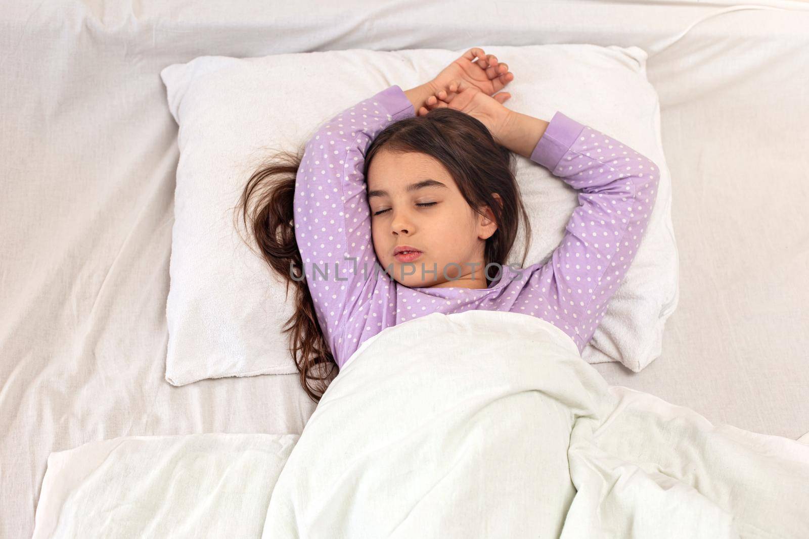 Little charming brunette girl with long hair in purple pajamas, sleeps lying on a white pillow, under a white blanket on the bed. View from above. Top view. Copy space