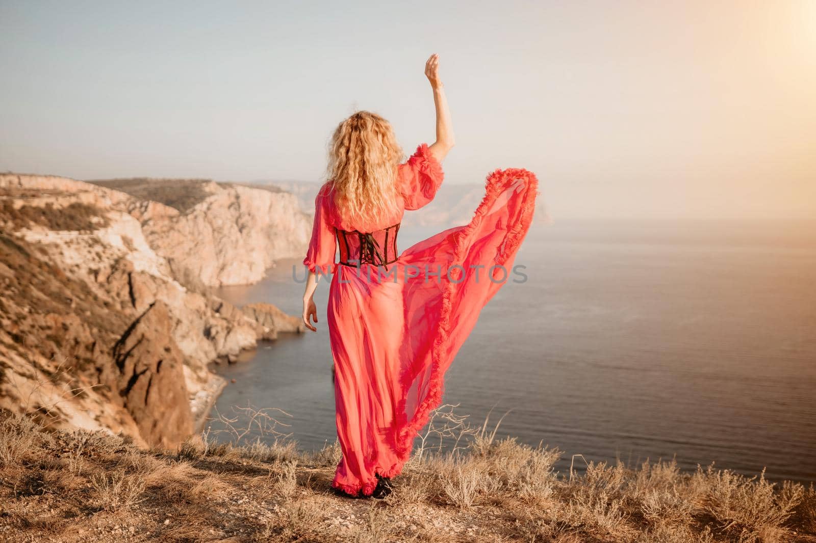 Side view a Young beautiful sensual woman in a red long dress posing on a volcanic rock high above the sea during sunset. Girl on the nature on blue sky background. Fashion photo