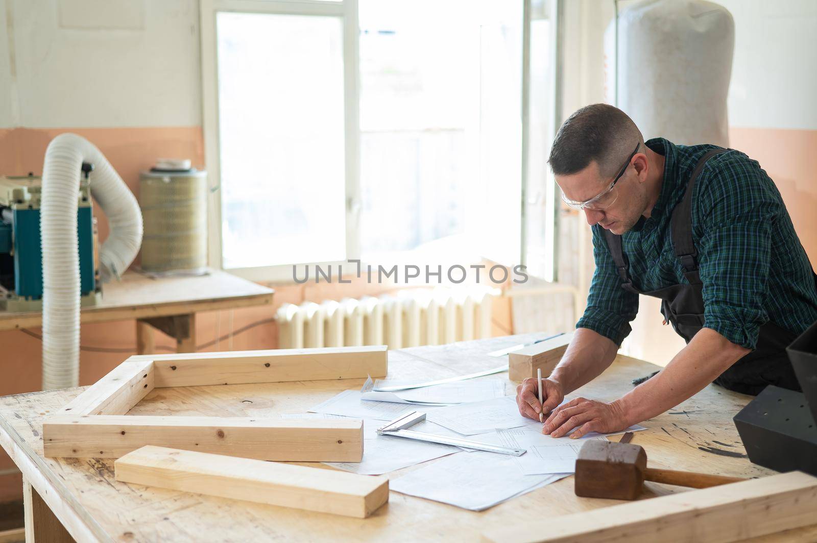 A portrait of a carpenter in goggles and work overalls draws a blueprint for a workshop. by mrwed54