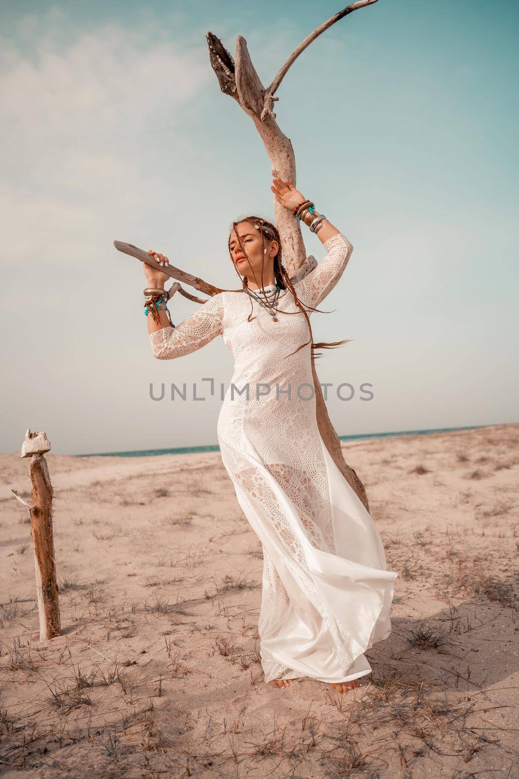 Model in boho style in a white long dress and silver jewelry on the beach. Her hair is braided, and there are many bracelets on her arms. by Matiunina