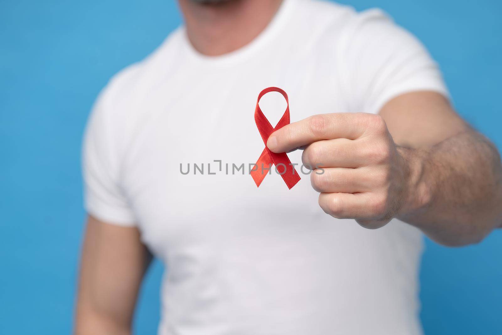 Man with red ribbon bow AIDS awareness symbol in hand wearing a white t-shirt isolated on a blue background. Modern medicine and healthcare. AIDS awareness concept. No face visible by LipikStockMedia