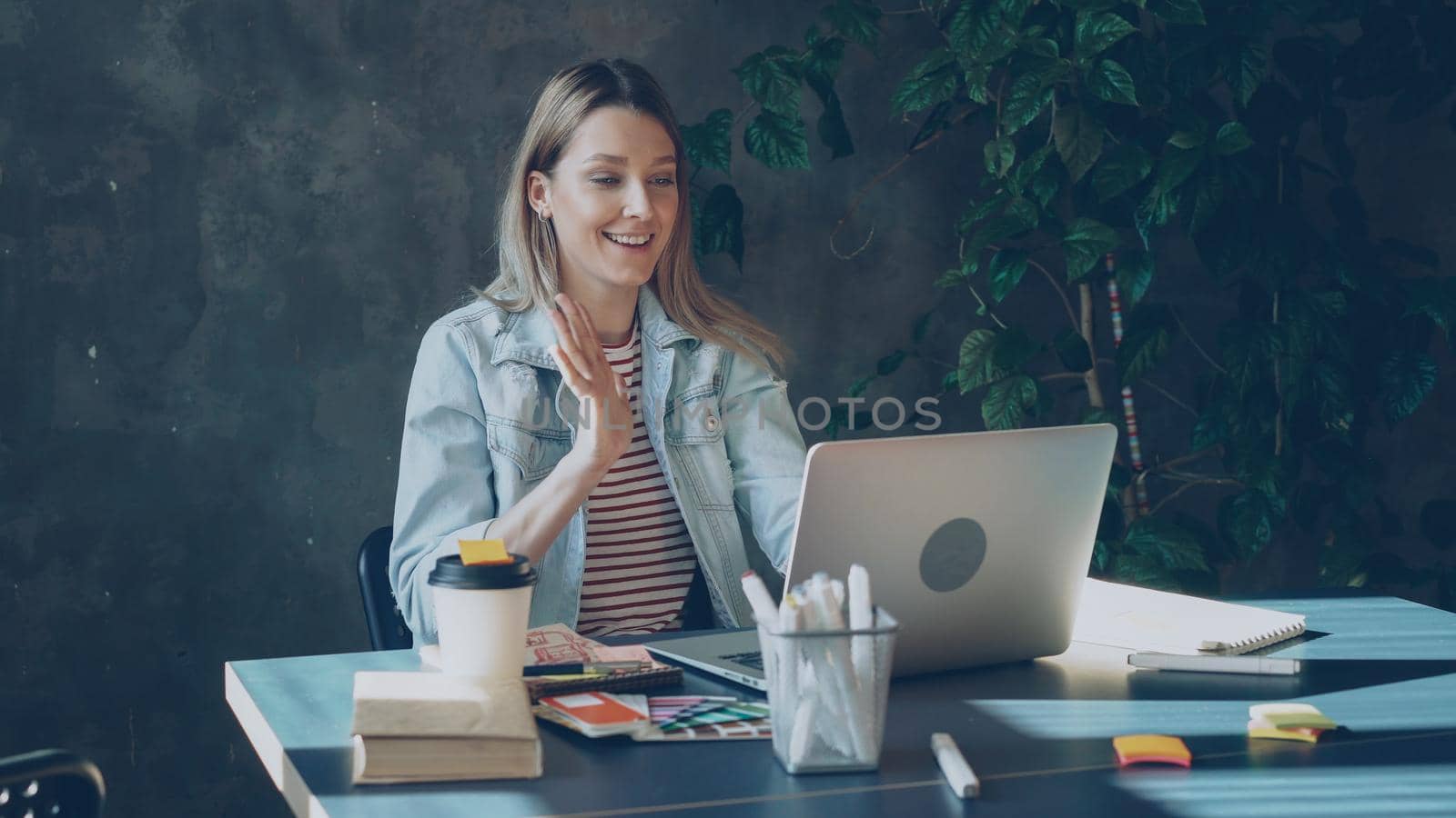 Attractive young woman is talking on skype on laptop while sitting at table in modern office. She is speaking emotionally, gesturing and laughing. by silverkblack