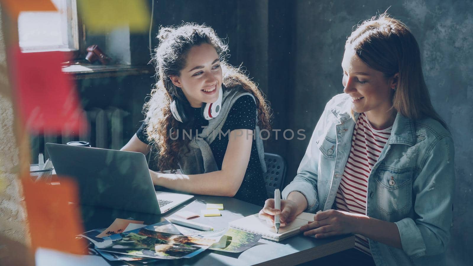 Cheerful pretty girl is working with laptop and listening to music, then taking off wireless headphones and talking with her coworker and laughing. Productive teamwork concept.