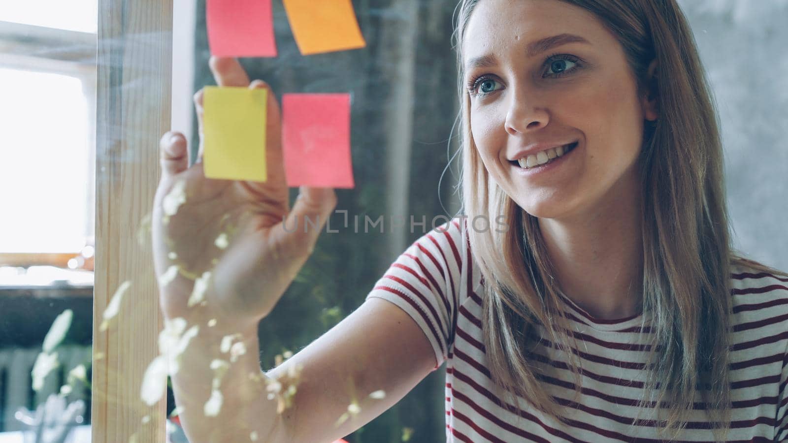 Close-up of pretty young woman with blond hair sticking colored memos on glass board in modern office. She is looking at papers and smiling. by silverkblack