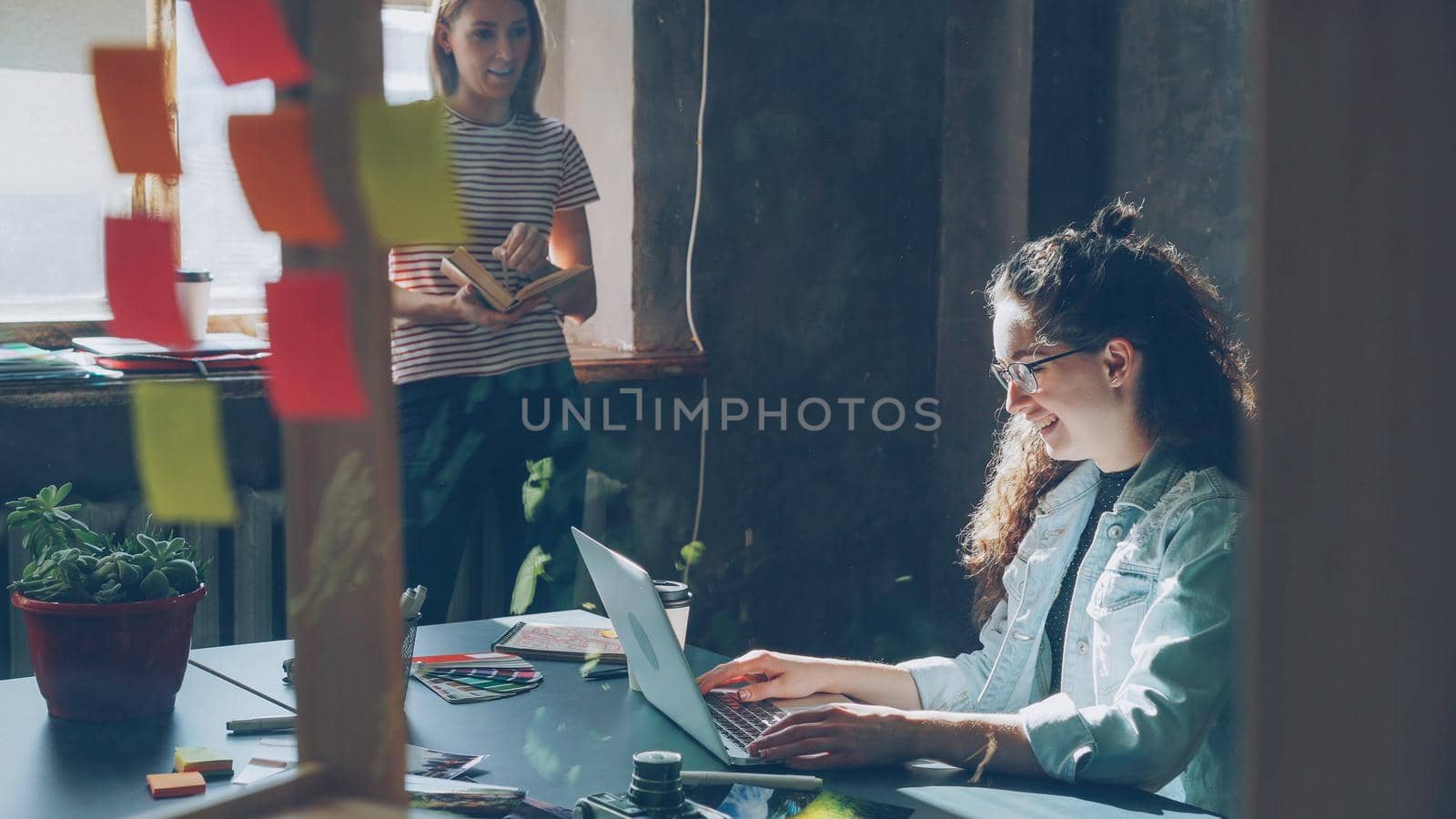 Two attractive girls are working together in modern loft office. Curly businesswoman is typing on laptop sitting at table while her colleague is reading book standing near window by silverkblack