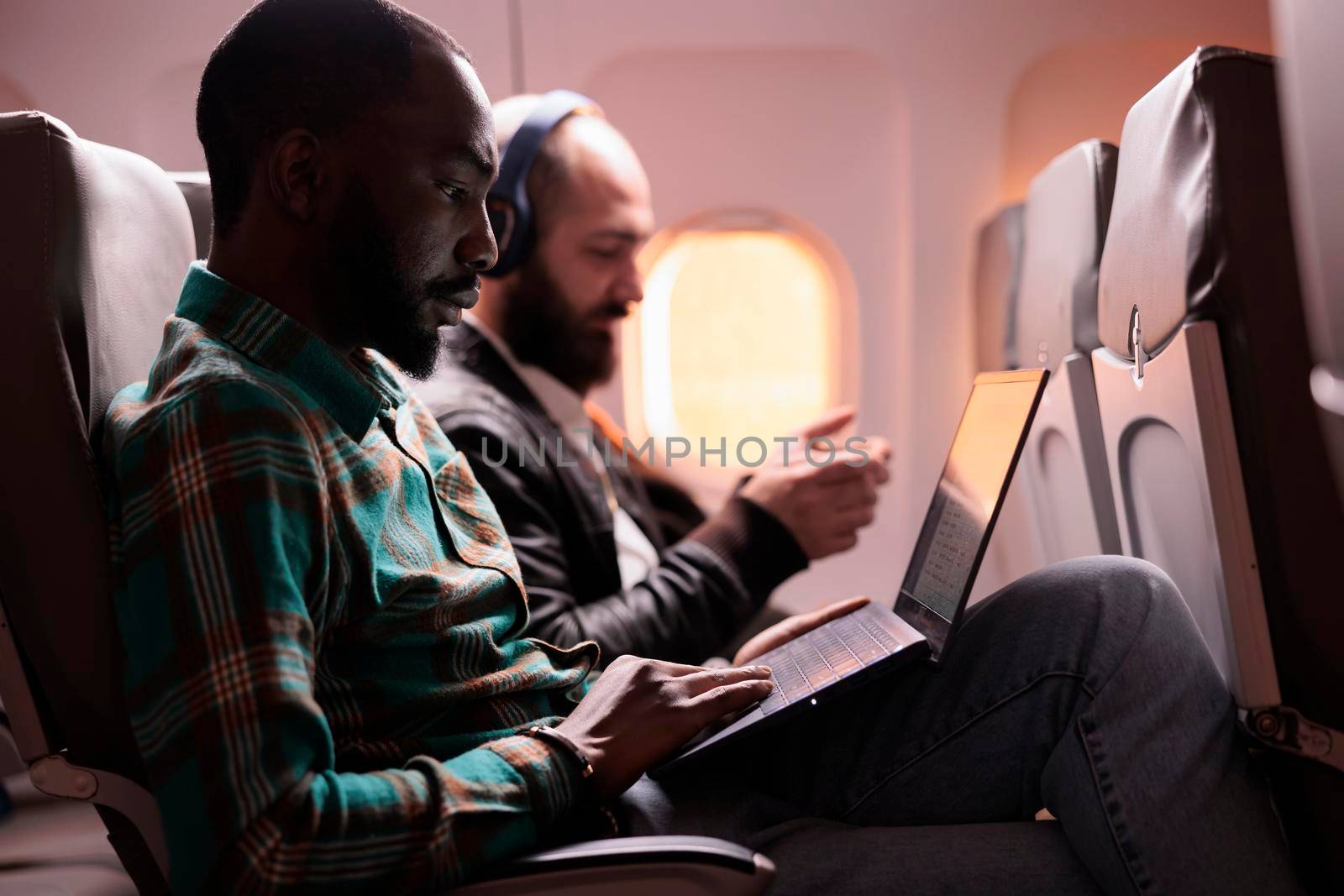 Businessman in seat travelling on work trip by airplane in economy class, working on laptop during sunset flight. Male employee flying with commercial flight with international airline.