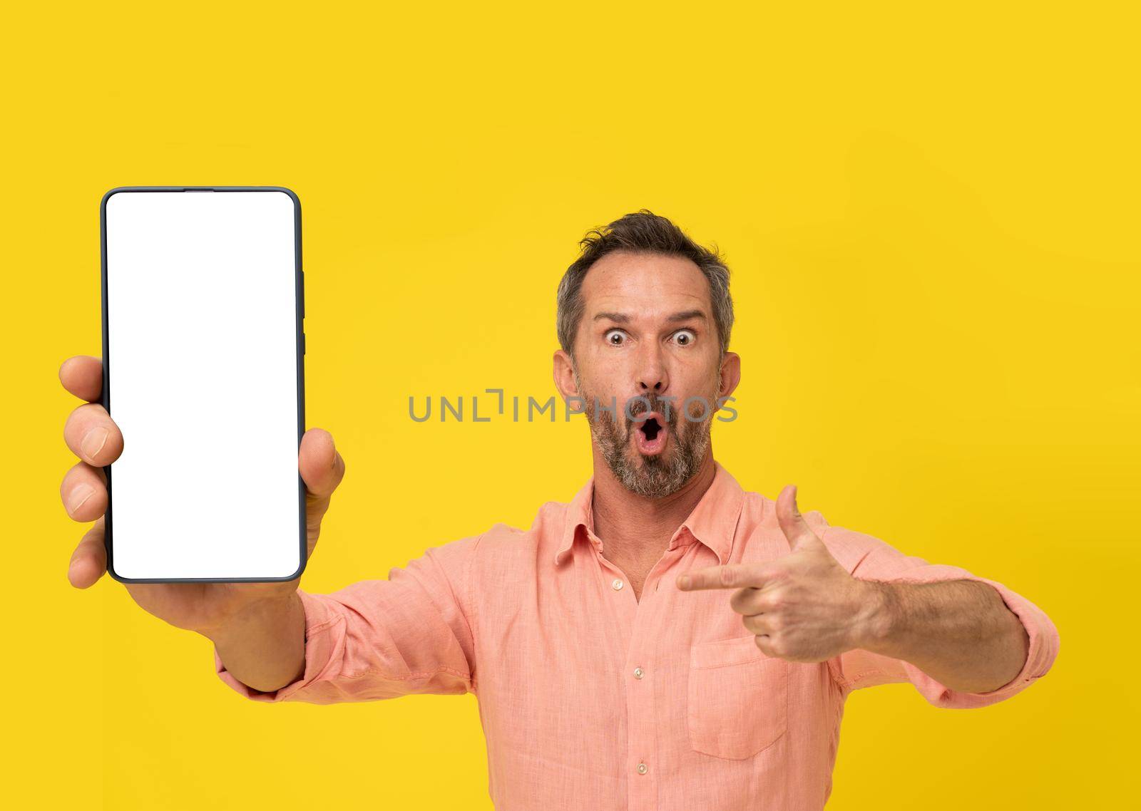 Amazed middle aged grey haired man pointing finger at smartphone looking at camera wearing peach shirt isolated on yellow. Mature fit man with phone app advertisement. Mock up white screen by LipikStockMedia