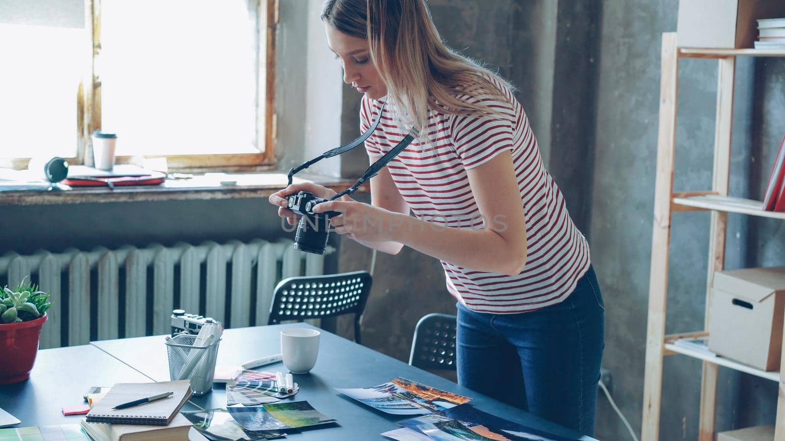 Young blond female photographer is making flat lay from colorful photos on table and shooting them with camera. She is displaying pictures near color palette and markers on desk.