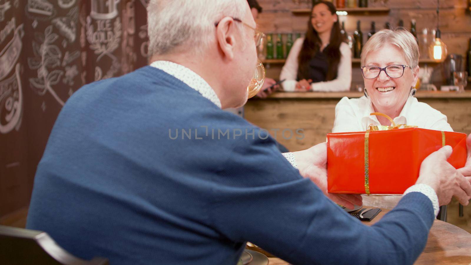 Senior man giving her wife an retirement gift during dinner in a restaurant by DCStudio