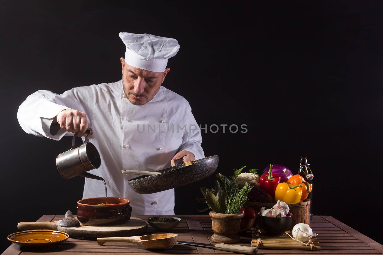 Male chef in white uniform and hat pouring olive oil onto cooking pan with vegetables by bepsimage