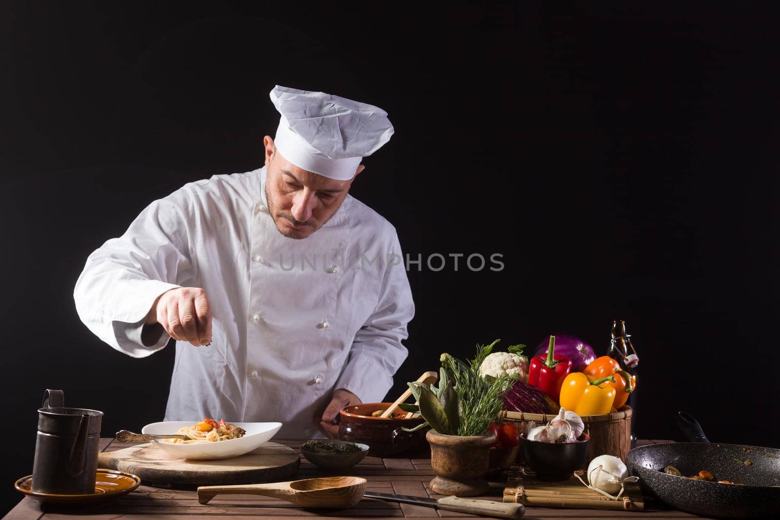 Male cook in white uniform and hat putting pot herbs on on food plate with spaghetti and vegetable before serving while working in a restaurant kitchen