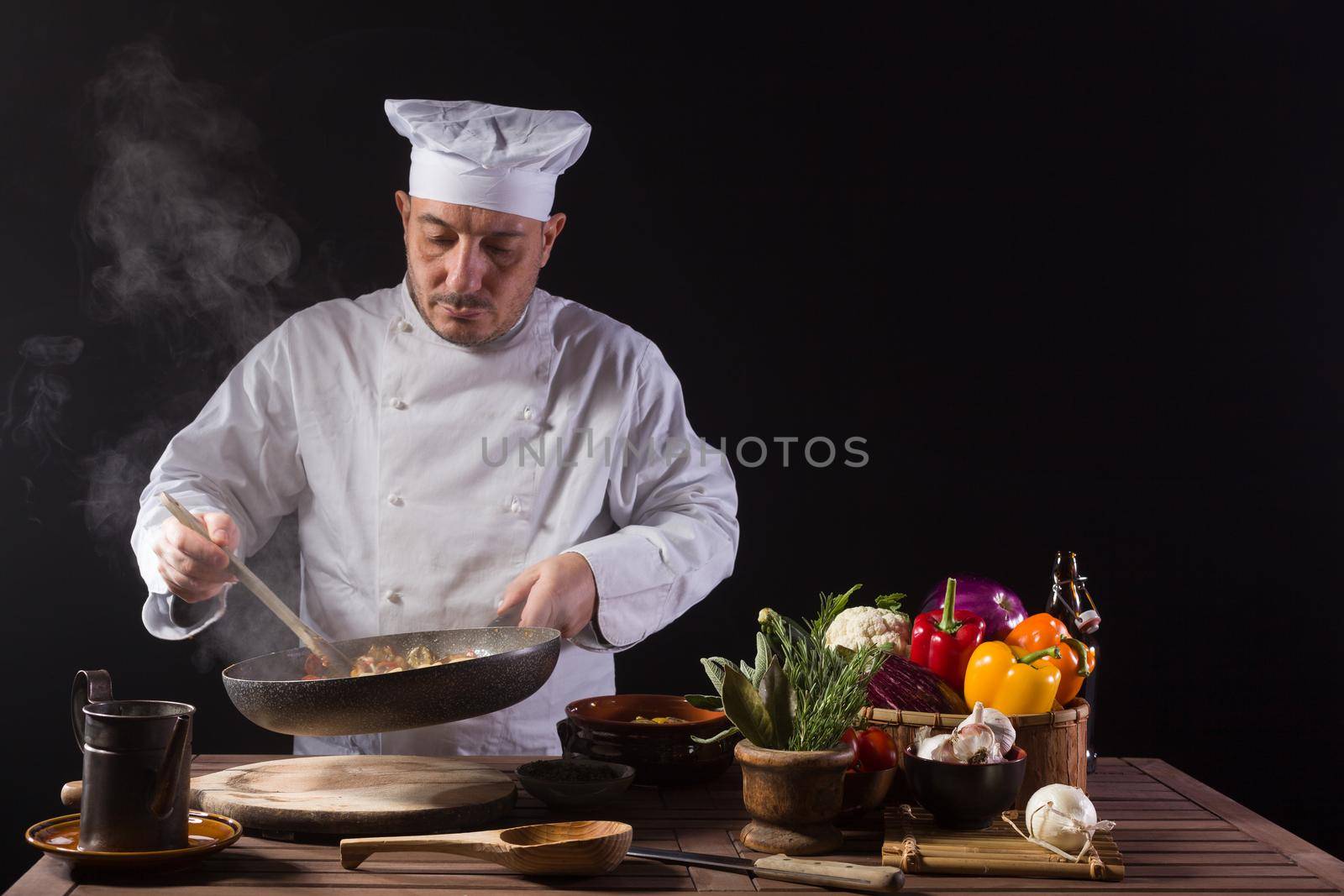 Male cook in white uniform and hat with ladle mixes the ingredients onto the cooking pan before serving while working in a restaurant kitchen