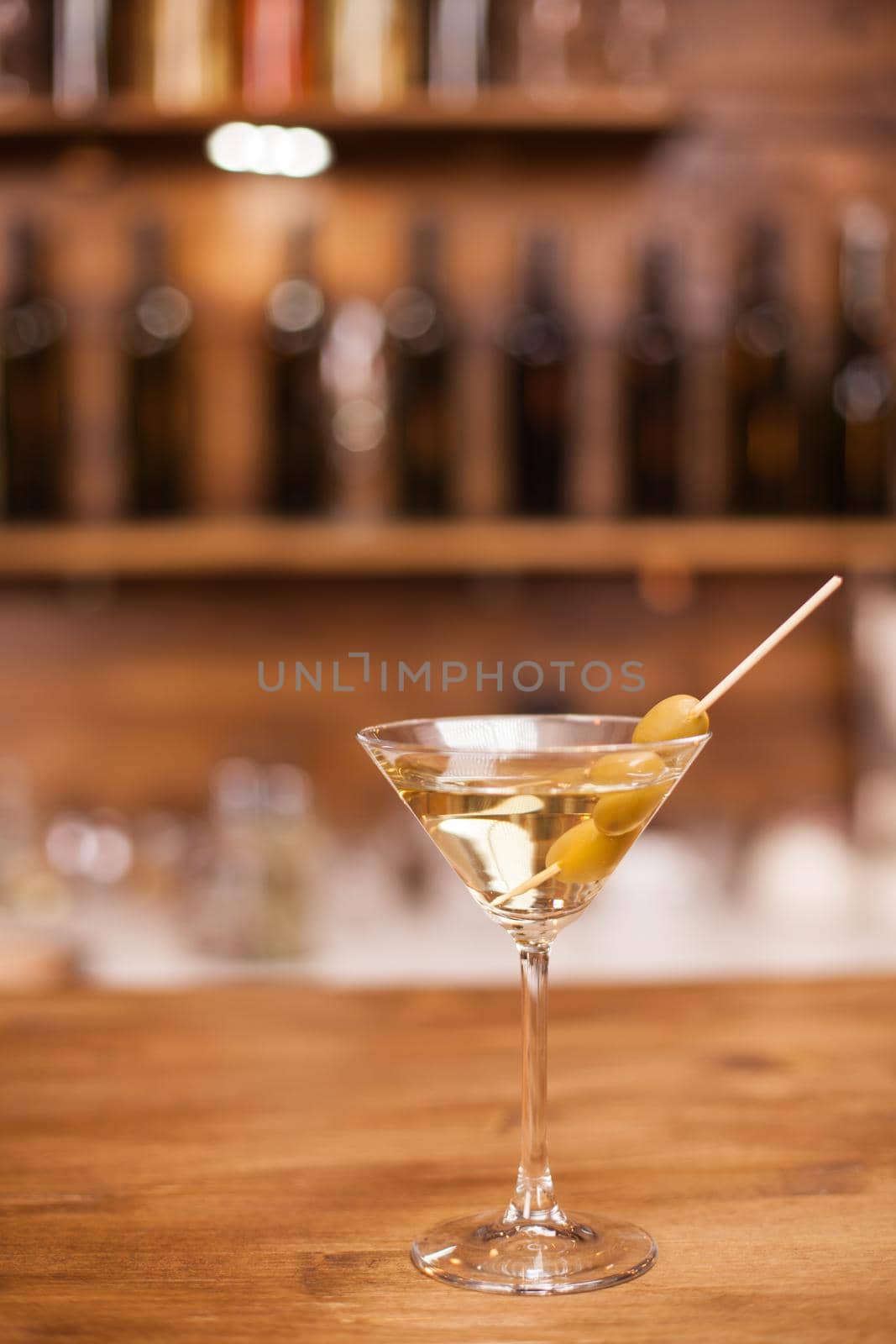 Glass of dry martini with green olives on a rustic wooden bar counter. Refreshment drink. Nightlife. Closeup