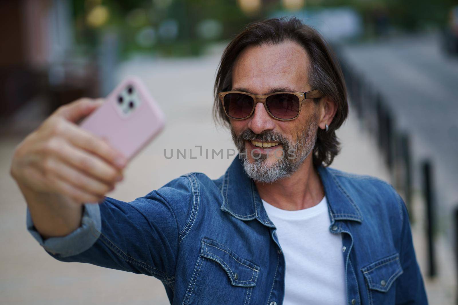 Man with smartphone outdoors making selfie or recording video message wearing denim shirt and sunglasses. Handsome middle aged man with grey hair making photos while traveling by LipikStockMedia