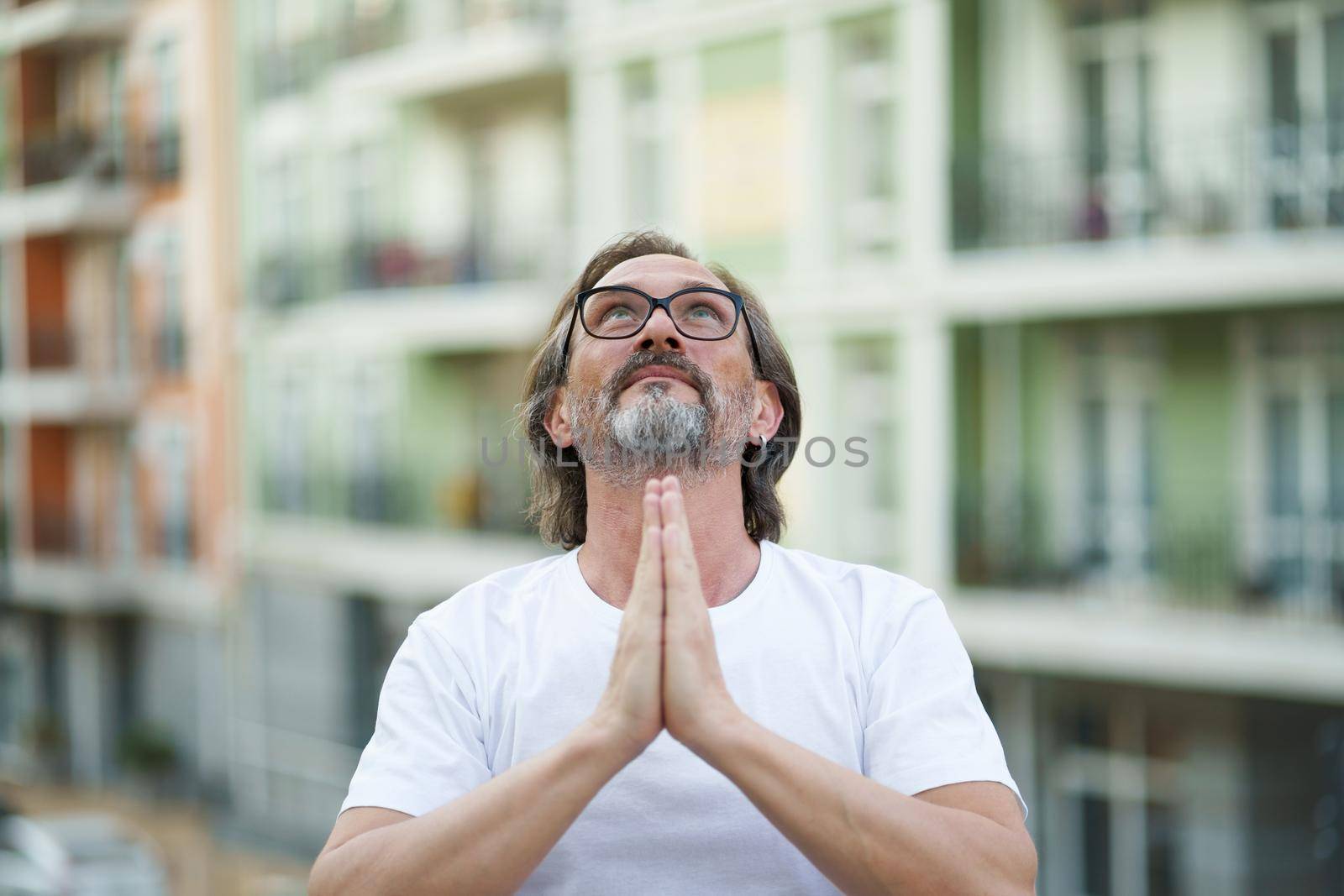 Giving praise to God put hands together mature man in glasses smile standing outdoors wearing white t-shirt. Happy mature man in eye glasses and looking up in prayer outdoor on a summer day.