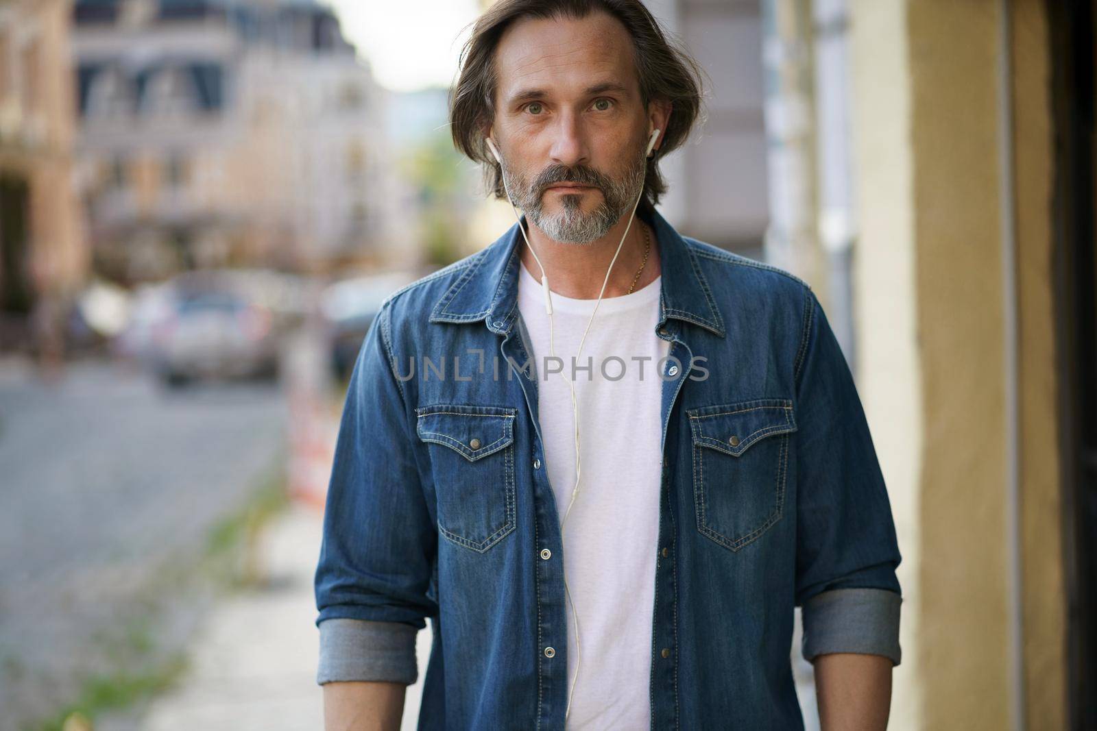 Handsome middle aged grey bearded man walking downtown with earphones listening music or talking on the phone wearing casual jeans shirt with white t-shirt. Travel lifestyle concept.